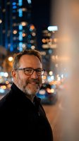 Smiling middle-aged Caucasian businessman with glasses and beard in urban night setting with bokeh city lights background for corporate portraits.