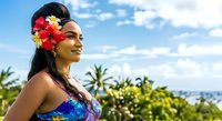 Young woman with dark hair and medium skin tone wearing tropical flower crown and colorful swimwear, smiling outdoors with palm trees and ocean backdrop for travel and summer campaigns.
