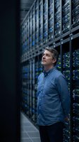 Middle-aged Caucasian IT administrator in blue shirt standing in modern data center server room with glowing LED lights for technology concepts.
