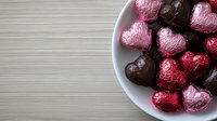Heart-shaped chocolate candies wrapped in pink and brown foil arranged on white ceramic plate against light wooden surface, romantic holiday confectionery.