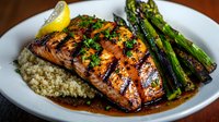 Grilled salmon fillet glazed with honey-soy sauce served on quinoa with charred asparagus and lemon garnish, drizzled with savory sauce on white plate.