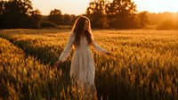 Young woman with long brown hair in white dress walking through golden wheat field at sunset with warm sunlight and peaceful rural countryside atmosphere.