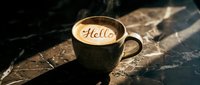 Steaming latte art coffee cup with Hello inscription on foam, placed on dark marble surface in warm morning light.