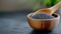Black poppy seeds in wooden bowl and spoon on dark background with green plant, selective focus, macro food photography for healthy eating concept.
