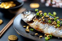 Grilled whole fish seasoned with soy sauce, garnished with green onions and roasted peanuts served on black ceramic plate with chopsticks, Asian cuisine.