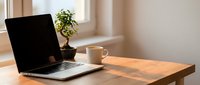 Laptop with blank screen on wooden desk next to coffee cup and potted plant by window, workspace setup for remote work or business projects.