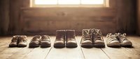 Row of various children shoes lined up on wooden floor in warm sunlight streaming through window creating nostalgic family moment atmosphere.