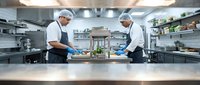 Two professional chefs in white uniforms and caps working together in modern commercial kitchen preparing food for restaurant service.