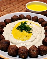 Traditional Middle Eastern hummus with falafel balls arranged around creamy dip topped with olive oil, herbs and spices on white plate, served with additional oil.