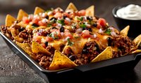 Loaded nachos topped with seasoned ground beef, melted cheese, fresh tomatoes, and chopped herbs served on black platter with sour cream dip.