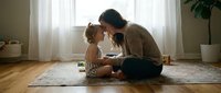 Mother and toddler daughter sharing tender moment reading book together on living room floor. Family bonding time with natural window light.