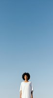 Woman with curly dark hair and medium skin tone wearing white casual shirt standing against clear blue sky, looking away thoughtfully for lifestyle and portrait use.