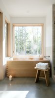Minimalist bathroom with wooden soaking tub positioned beneath large window overlooking forest landscape, featuring natural light, white towels on wooden stool, and concrete flooring for spa-like relaxation.