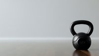 Black kettlebell weight on wooden floor against white wall in minimalist gym setting for fitness and strength training workouts.
