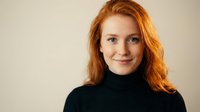 Young redhead woman with wavy hair wearing black turtleneck sweater smiling at camera against neutral beige background for professional headshot.