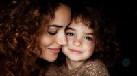 Young Hispanic mother and daughter with matching curly brown hair share tender moment, faces close together, expressing love and bonding against dark background.