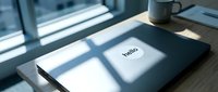 Laptop with hello sticker on desk by sunny window, coffee mug nearby, modern workspace with natural light and clean minimalist style.