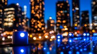Glowing blue lights illuminate urban waterfront with blurred city skyline and bokeh reflections creating atmospheric nighttime cityscape scene.