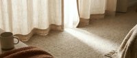 Sunlit bedroom corner with beige carpet, flowing linen curtains, ceramic mug, and cozy knit blanket creating a serene, minimalist morning atmosphere for home decor and lifestyle content.