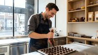 Male artisan in apron crafting handmade chocolates in bright studio kitchen, arranging confections on tray near window with shelving, professional food production workspace.