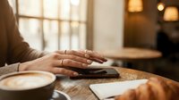 Woman's hands typing on laptop keyboard at wooden table in warm cafe setting with natural window light and cozy atmosphere for remote work and productivity.