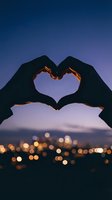 Hands forming heart shape silhouette against twilight sky with city lights bokeh background for romantic concepts and love themes.