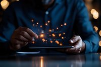 Male hands holding digital tablet with glowing orange particles floating above screen, creating magical atmosphere against dark background with bokeh lights.