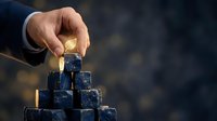 Caucasian male hand placing gold coin on top of pyramid stack of dark blue cubes, symbolizing investment growth, financial success, and wealth accumulation strategy.