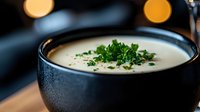 Creamy soup with fresh herbs in dark ceramic bowl on wooden table with warm bokeh lights creating cozy restaurant atmosphere for comfort food dining.
