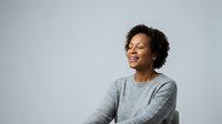Middle-aged woman with curly dark hair and medium brown skin tone wearing gray sweater, smiling while looking to the side against white background for portrait and lifestyle use.