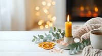 Cozy winter home interior with burning candle, eucalyptus branch, dried orange slices and warm knitted blanket creating peaceful hygge atmosphere.