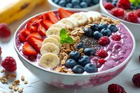 Healthy smoothie bowl topped with fresh strawberries, bananas, blueberries, raspberries, granola, chia seeds and mint leaf garnish on white marble background.