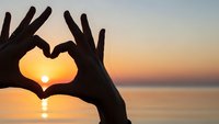Hands forming heart shape silhouette against golden sunset over calm ocean water creating romantic peaceful moment for love and connection concepts.