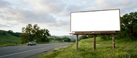 Large blank white billboard on wooden posts beside rural highway with green hills and trees under cloudy sky for advertising campaigns.