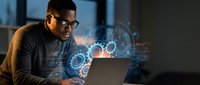 Young African American male software engineer working late on laptop with holographic gear interface projections in modern office environment.
