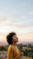 Young woman with curly dark hair and medium brown skin tone wearing mustard yellow ribbed sweater, smiling while looking upward at scenic sunset landscape with rolling hills and trees below.