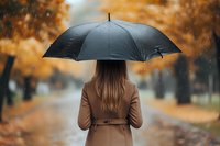 Woman in beige coat holding black umbrella standing on autumn alley with falling rain and orange foliage in blurred background, rear view atmospheric photo.