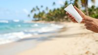 Close-up of hand holding white sunscreen bottle on tropical beach with turquoise ocean and palm trees, ideal for skincare and sun protection marketing.