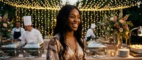 Young African American woman smiling at elegant outdoor dinner party with string lights and chefs preparing food in background for luxury dining events.
