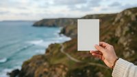 Caucasian hand holding blank white card against coastal cliffs and turquoise ocean, ideal for mockup and travel marketing campaigns.