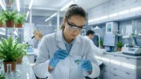 Asian female scientist in lab coat and safety glasses examining test tube sample in modern research laboratory with plants and equipment.
