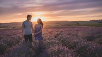Couple standing together in blooming lavender field at golden hour sunset, romantic outdoor engagement or relationship portrait for lifestyle and travel content.
