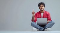 Young South Asian man in red shirt sitting cross-legged with laptop, pointing upward with idea gesture against gray background.