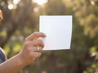 Person holding blank white card outdoors in natural sunlight with blurred green foliage background, suitable for mockups, advertising, and promotional designs.