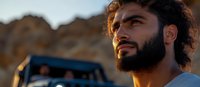 Bearded man with curly dark hair and Middle Eastern ethnicity gazes thoughtfully into distance during golden hour sunset, with off-road vehicle and desert landscape behind.