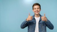 Happy teenage boy giving thumbs up gesture wearing denim jacket on blue background. Positive young male student showing approval and success.