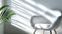 Modern gray armchair with wooden legs against white wall with dramatic window light and shadow patterns, green palm leaf adds natural accent. Perfect for interior design.
