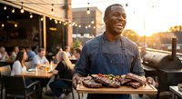 Happy African American chef presenting grilled meat platter at outdoor restaurant during golden hour with string lights and dining guests in background.