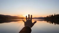 Hand silhouette reaching toward golden sunset over calm lake with mountain reflections creating peaceful moment of connection with nature.