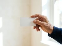 Mature man with medium skin tone holding blank white card in hand against bright window background, suitable for business communication, greeting cards, and corporate messaging concepts.